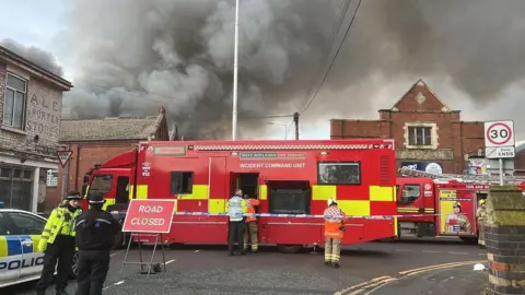 A big red vehicle with "incident command unit" on the side is behind a strip of police blue and white tape with a "road closed" sign in front of it. Several firefighters and police officers can be seen in front of the vehicle with a huge smoke plume behind.