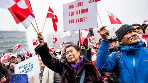 Getty Images A crowd of protesters holding red and white Greenlandic flags and signs rally at City Square in Copenhagen, Denmark. They are dressed in winter coats, with one man wearing a beanie. A sign at the top of the image reads: "Our land, our freedom, our voice!"