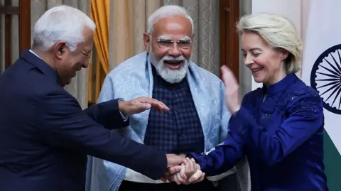 Reuters Three leaders are standing close together indoors, smiling and laughing during a friendly moment. The man in the centre, Indian Prime Minister Narendra Modi, with a white beard and glasses, wears a light shawl over a dark outfit and appears animated. On either side, European Council President Ant&oacute;nio Lu&iacute;s Santos da Costa (left) in a dark suit and European Commission President Ursula von der Leyen (right) a deep blue jacket lean in, clasping hands in a gesture of warmth. A national flag and curtains are visible behind them