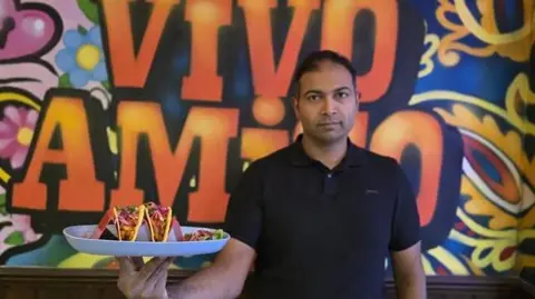 Rajendra Vikram Kupperi Rajendra Vikram Kupperi wearing a black short-sleeved shirt is holding a large white plate with three vibrant tacos. Each taco is filled with colorful ingredients, including shredded lettuce, diced tomatoes, and possibly meat, topped with bright pink pickled onions. The background features a bold, colorful mural with swirling patterns and large orange letters spelling &ldquo;VIVO AMIGO&rdquo; against a mix of blue, yellow, and pink designs.
