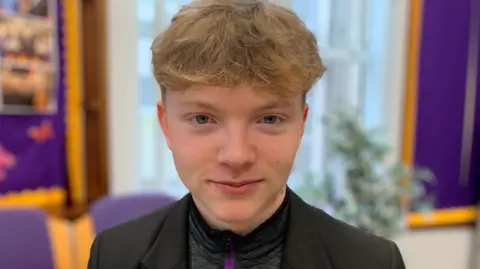 Matthew has fair hair.. He is wearing a black blazer over a yellow, purple and grey zip up top. The blurry background shows purple walls.
