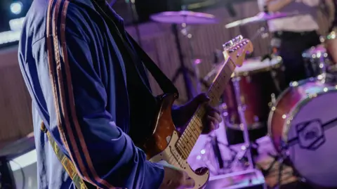An unidentified man in a blue jacket plays a guitar on a stage with a drum set in the background.