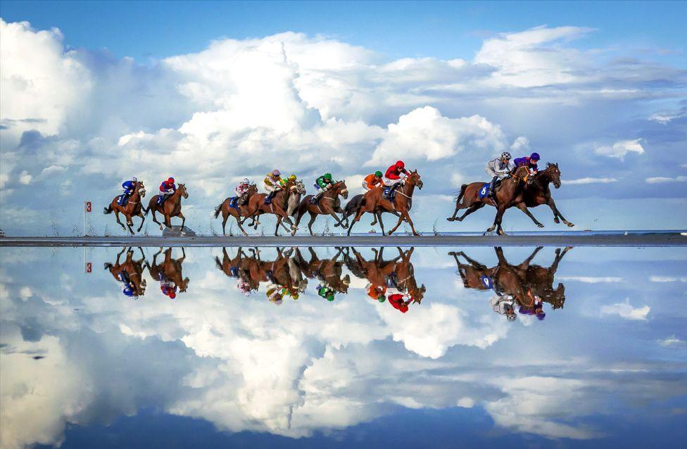 A group of jockeys riding horses at full speed on a wet track, creating a vivid reflection of the horses and riders in the water. The race takes place in Laytown, Ireland's only horse racing event run on a beach under the Rules of Racing