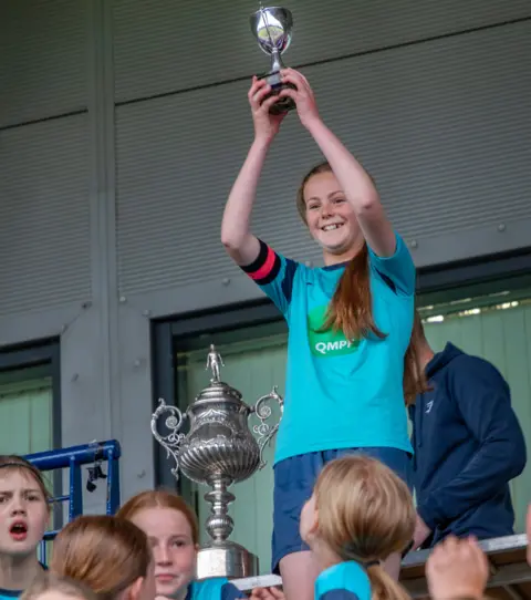 Stephen Tierney Clara has long brown hair in a ponytail. She is wearing a light blue football shirt. There is a large trophy beside her. There are children in the foreground.