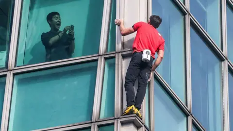 Getty Images A building occupant uses his phone to record US rock climber Alex Honnold climbing the Taipei 101 building. Honnold is wearing a red t-shirt and black trousers with yellow shoes.