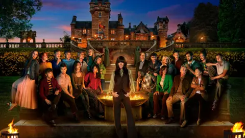 BBC/PA The contestants taking part in this year's The Traitors sit outside the castle at night time near a large fire pit. Claudia Winkleman is standing in front of the fire place looking directly at the camera. In the background is a large castle against the setting sun.
