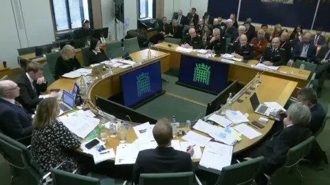 House of Commons Chief Constable Craig Guildford sitting before the select committee at the House of Commons. He is flanked by other officers facing eight MPs around an oval table.
