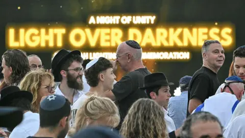 Getty Images Mourners arrive to attend the memorial held for the victims of a shooting at Bondi Beach. A screen reading, "a night of unity", "light over darkness" can be seen in the background.