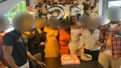 BBC A group of 11 young people stand close together next to a cake in the shape of the number 21. All their faces are blurred apart from that of a young woman smiling in the middle.