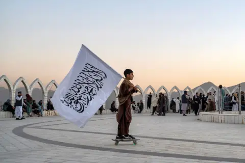 AFP via Getty Images An Afghan boy carries the Taliban flag as he skates across the Wazir Akbar Khan hilltop in Kabul on October 13, 2025.