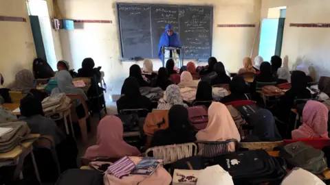 Schoolgirls are sitting at desks in a classroom looking at their teacher, a woman, who is standing in front of a blackboard.