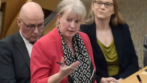 PA Media Shona Robison, who has blonde hair tied back, speaks in the Scottish Parliament chamber with her right hand held out in front of her, holding a pen. She is wearing a pink suit jacket and a black and white scarf.