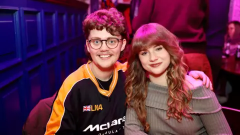 Jonathan Phang Kayleb and Alana smile at the camera in a pub. Kayleb has short curly hair and wears a racing jersey. Alana has long curled hair and wears a beige jumper.