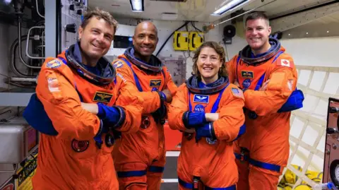 NASA Four astronauts wearing bright orange spacesuits but no helmets. From left to right they are Reid Wiseman, Victor Glover, Christina Koch and Jeremy Hansen. They are standing together positioned in a row with their arms crossed. The room they are in is filled with technical equipment and cabling.