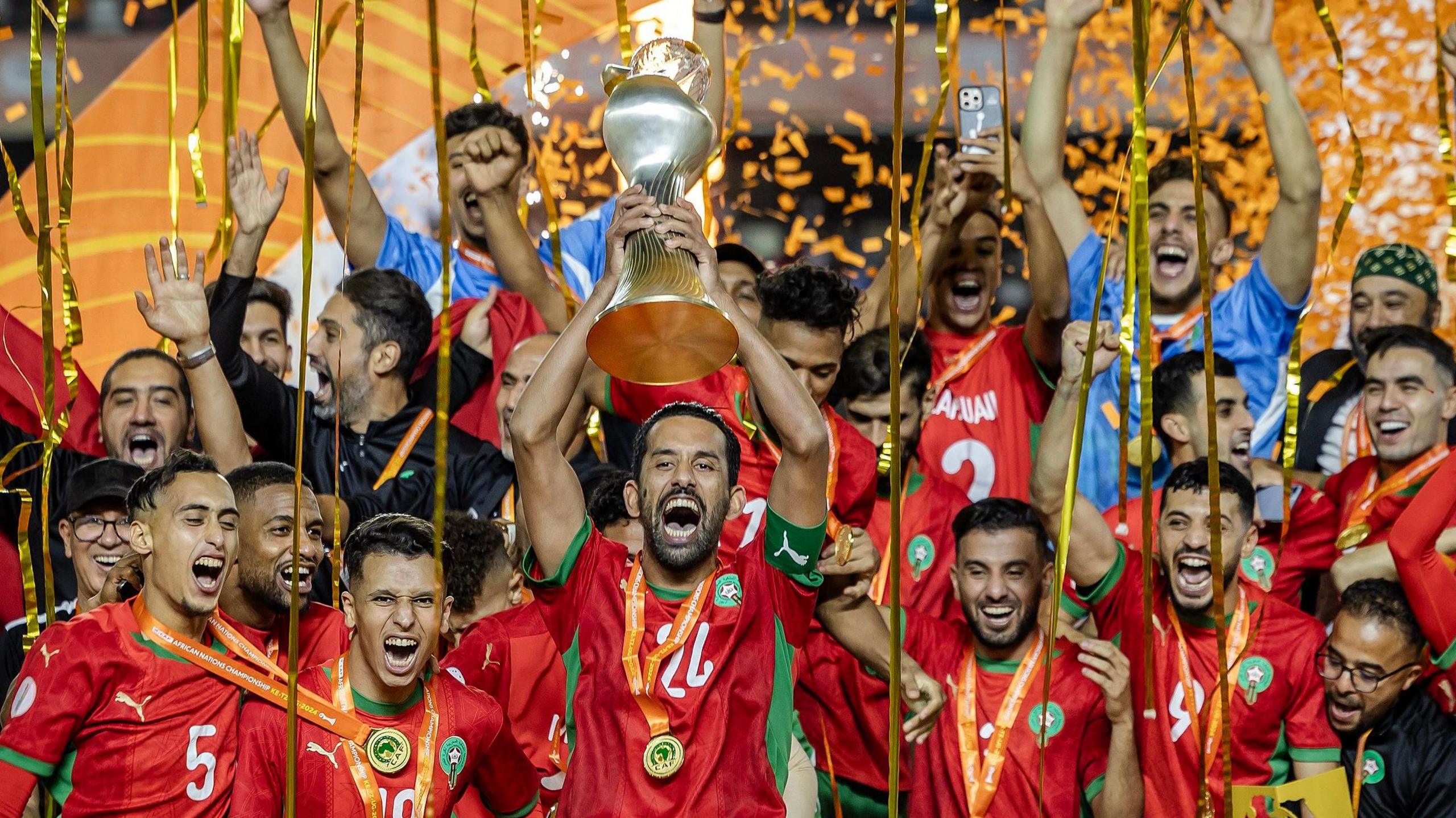 Morocco captain Mohamed Rabie Hrimat holds the curved, vase-like African Nations Championship trophy aloft as he is surrounded by team-mates in red shirts with medals around their necks and streamers of orange ticker tape coming down around them