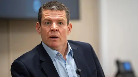 PA Media Rhun ap Iorwerth sat giving a press conference with an open necked shirt and dark blue jacket.