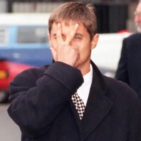 PA Media A younger John Alford makes a V-sign in front of his face with his right hand. He is wearing a smart, heavy coat, white shirt and checked tie.