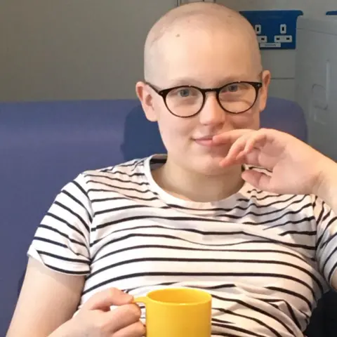 Louise Dempster A young girl wearing dark-rimmed glasses and a black and white striped t-shirt. She is holding a yellow mug and sitting on a blue sofa. She has no hair.