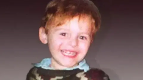 PA Media Toddler James Bulger is wearing a brown jumper over a blue shirt and is smiling at the camera.