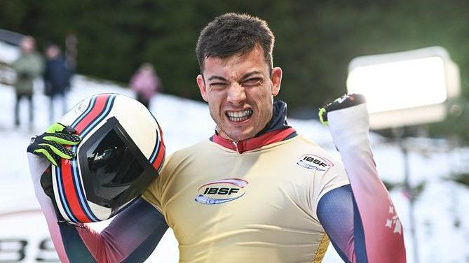 Matt Weston celebrates, looking into the camera and raising his left fist while holding his crash helmet in his other hand