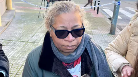 BBC/Moose Campbell A woman in a grey scarf and coat talks to reporters