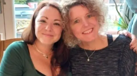 Family photo Helen and Rachael side by side smiling with their heads close together, with Helen's arm around Rachael's shoulder. One has curly blonde hair, and the other long dark hair.