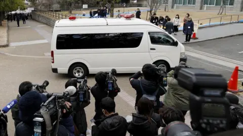 Reuters A white police van carrying Tetsuya Yamagami arrives at a Nara district court ahead of his sentencing hearing. Journalists and videographers are crowding around it