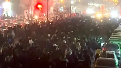 User generated content People viewed from behind march along a Tehran street at night, on 8 January 2026. A line of cars is to the right of the demonstrators, several lanes of cars to the left, with bright lights in the distance.