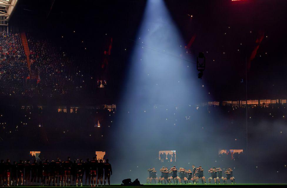 The All Blacks perform a synchronized haka under a dramatic spotlight on a dimly lit stadium field.