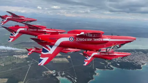 MOD Red Arrows bearing Union flag livery flying upside down in formation along a coastal strip with land in the background.