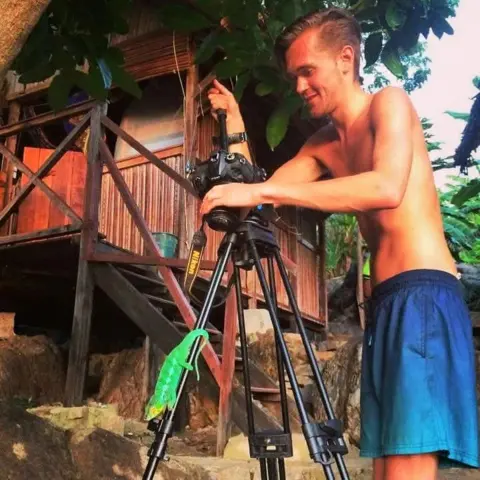 Christian Marot A man wearing swimming trunks is filming a lizard clinging to the leg of his tripod with jungle foliage and a wooden hut behind him