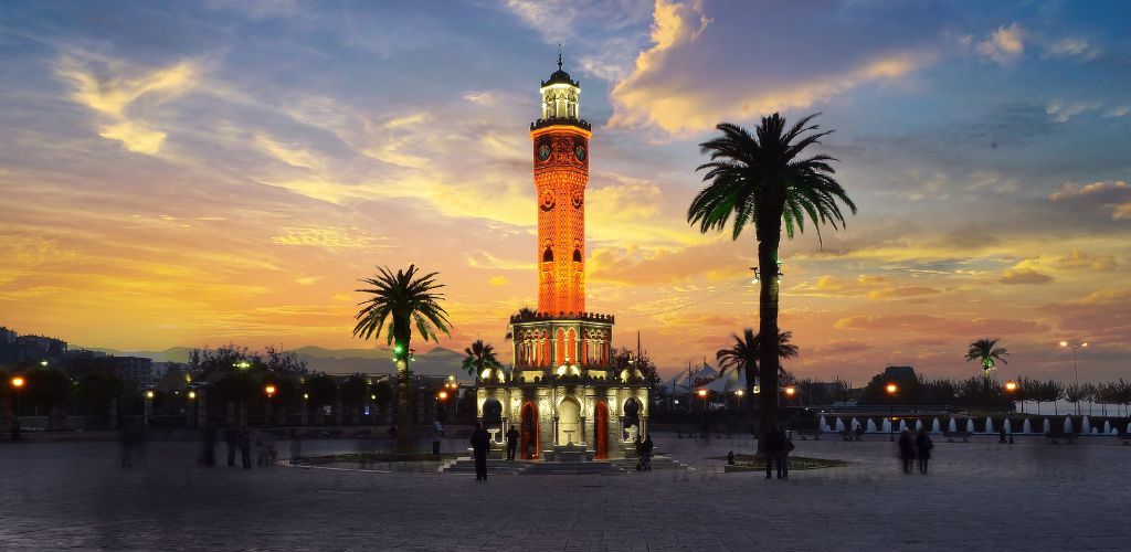 A tower of Izmir clock stands at the center of the park with trees and some people walking around.