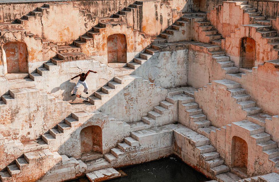 A skateboarder performs a trick mid-air among the geometric steps of an ancient stone stepwell. The weathered walls and repeating stair patterns create a striking architectural backdrop.