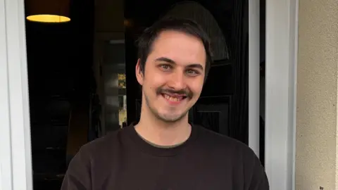 BEN SCHOFIELD/BBC Jake Pateman standing in the doorway of his house. He is smiling and looking directly down the camera. He is wearing a black sweatshirt and has dark hair and dark eyes. The door of his home is open, but inside it is too dark to make anything out, besides a sliver of a window in the distance.