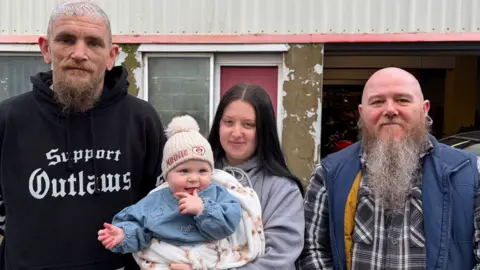 Alex standing in the middle holding Lilah in a cream blanket next to Wesley and Marc in front of a garage. They are all looking at the camera. Wesley, on the left, wears a black hoodie and is bald with tattoos on his head. He has a ginger beard. Alex is wearing a grey hoodie and has long black hair. Baby Lilah is smiling and has one of her hands near her mouth. On the right, Marc wears a grey tartan shirt and a blue gilet. He is bald with a long grey beard.