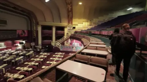 Urbandoned Two people walk along an aisle in what appears to be an abandoned theatres building. Red cushion seats can be seen across the site.