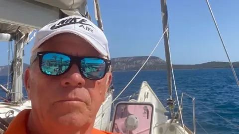 Laurie Watson Middle-aged man on a sailing boat faces the camera wearing black sunglasses with reflective lenses, an orange top and white cap