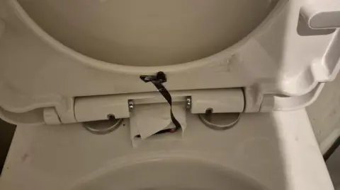 Supplied The lifted lid of a white toilet seat, with a black camera tucked under its rim and a wire leading to another part of it wrapped in toilet paper