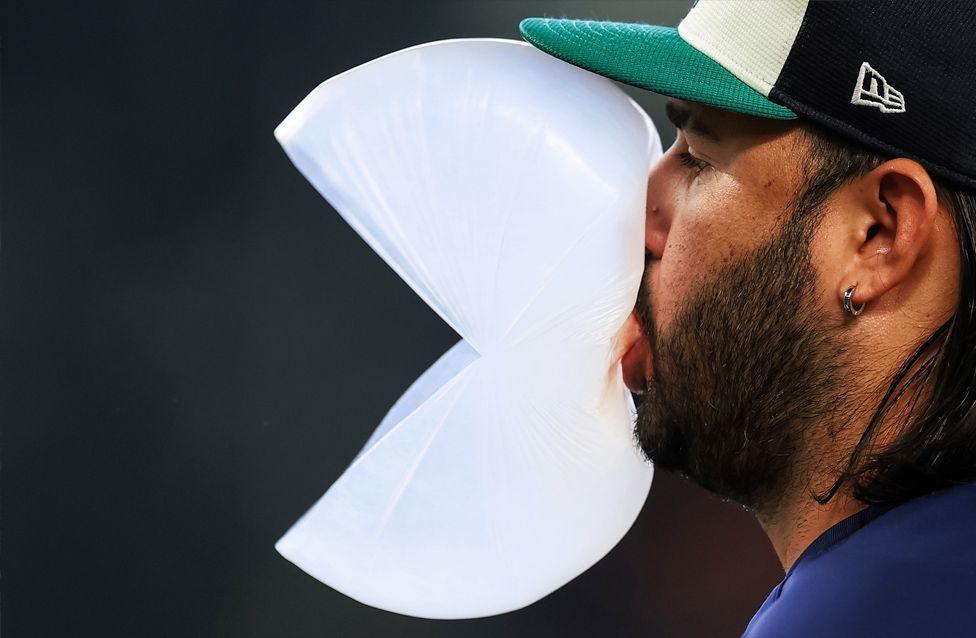 A close-up of baseball player Eugenio Suárez blowing a large bubble with gum, shaped like an open Pac-Man figure. The player wears a dark cap with a visible logo and an earring.
