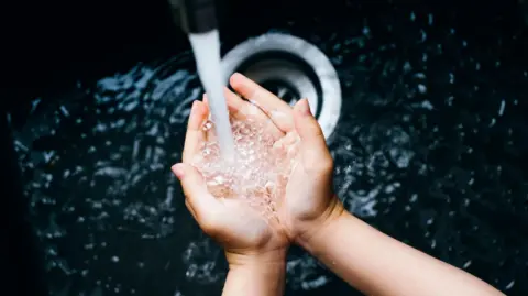 Getty Images A young woman's hands cup water as it runs from a tap into a deep black sink.