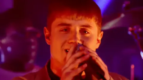 John Alford performing on Top of The Pops, holding a microphone to his mouth as he sings.