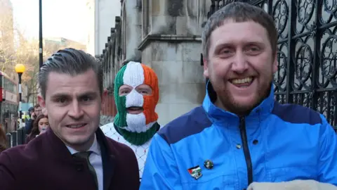 PA Media Lawyer Darragh Mackin wearing a maroon coat arrives with DJ Pr&oacute;va&iacute;, in a tricolour mask and white jumper and Dan Lambert in a blue coat.