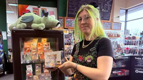 Amy Wosley Amy standing in a caf&eacute; surrounded by trading cards holding a Pok&eacute;mon box