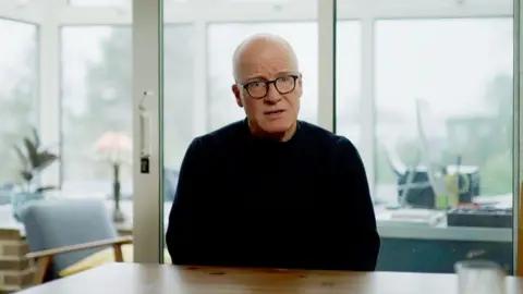 A bald man wearing black rimmed glasses and a black top is looking towards the camera. He is sitting at a wooden table. The background - a conservatory and garden - is blurred.