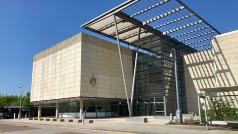 EMMA HOWGEGO/BBC The outside of the South Cambridgeshire District Council building. It is a concrete cube-shaped building supported by concrete columns with a glass building in between covered by a metal canopy.