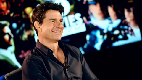 Cruise smiles as he looks off camera. He wears a black shirt. He has dark, black hair