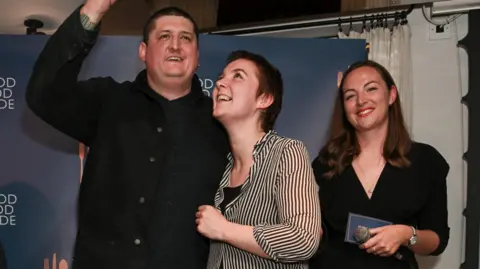 Getty Images Chef Gareth Ward stands on a small stage in front of a &ldquo;The Good Food Guide&rdquo; backdrop. He raises an award plaque above his head while smiling, his guest, a woman, looks up at it, smiling, and a woman on the right smiles while holding a microphone and a card.