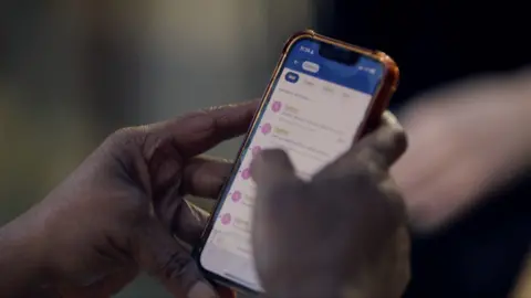 A woman's hands are holding a mobile phone, and her right-hand thumb is scrolling through emails. The background is blurred.