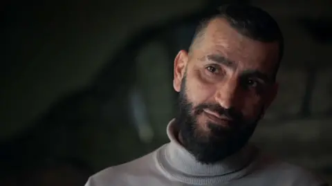 Habib in a white roll-neck top speaking to the camera against a dark background. He has a dark beard and moustache.