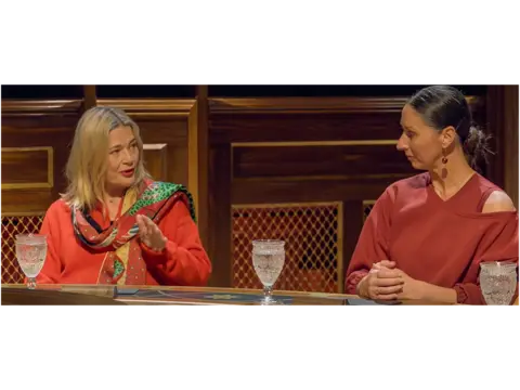 Harriet, in a red cardigan and red scarf, is sat next to Rachel, in a red dress, at the Traitors round table